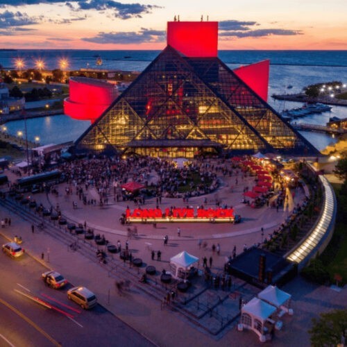 A large crowd gathers outside the glass pyramid-shaped Rock & Roll Hall of Fame in Cleveland at sunset, with red lighting illuminating the building and a "LONG LIVE ROCK" sign—where Warren Zanes once discussed Bruce Springsteen's enduring legacy.