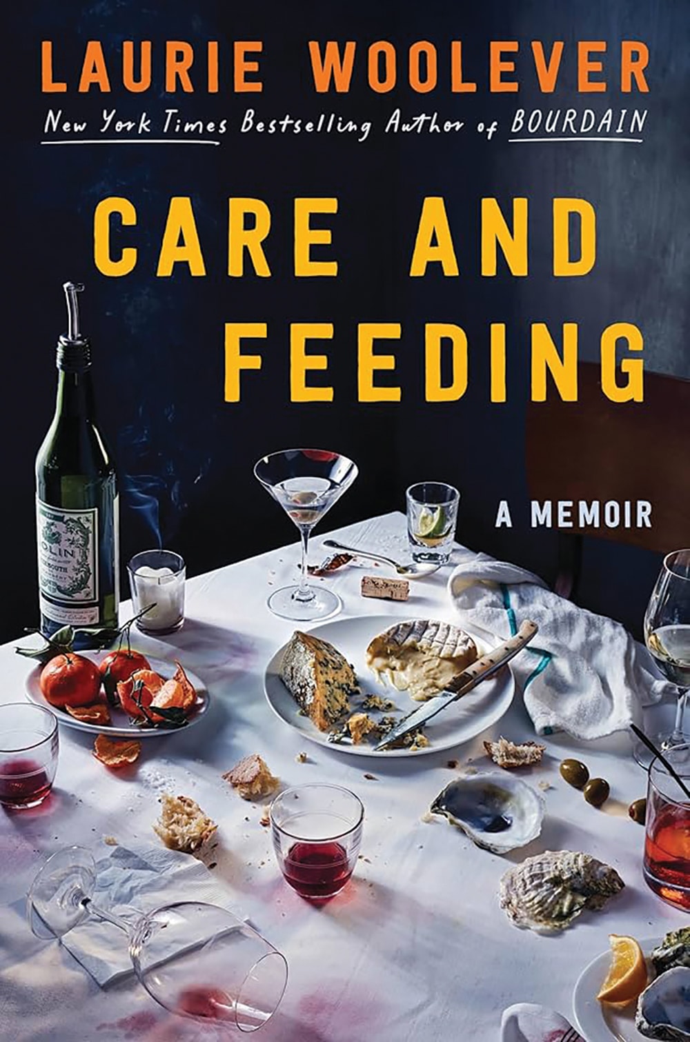 Book cover "Care and Feeding" by author Laurie Woolever A messy table after a meal with empty glasses, a wine bottle, half-eaten bread, cheese, and tomatoes evokes the spirit of Bourdain. Scattered utensils hint at stories.