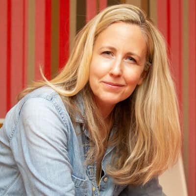 A woman with long blonde hair, wearing a light denim shirt, sits in front of a wall with vertical red, pink, and brown stripes, looking at the camera with a gentle expression.