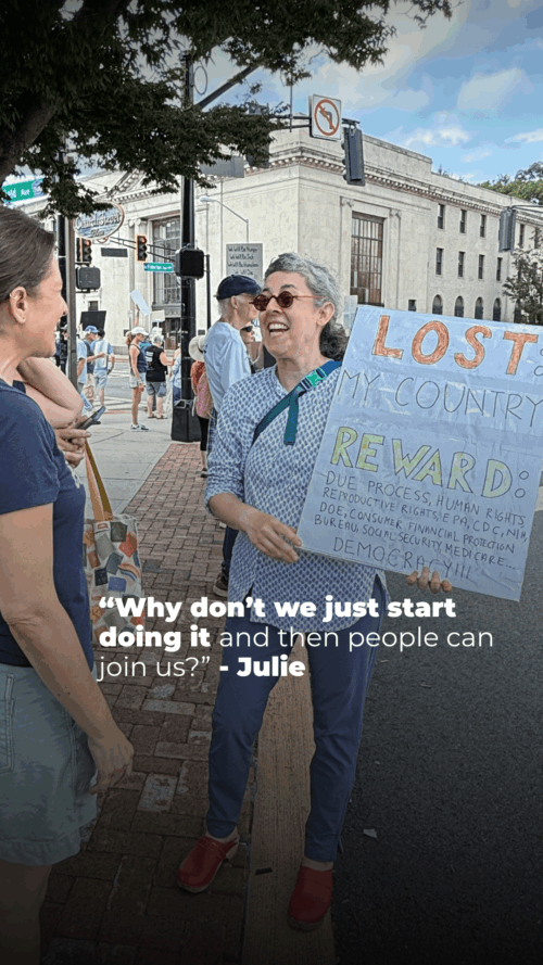 A smiling person with gray hair and glasses holds a large protest sign reading “LOST: MY COUNTRY” at a street demonstration, talking with another woman as people gather on the sidewalk in the background.