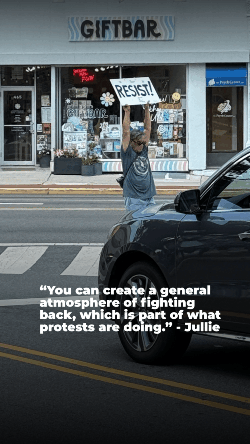 A person stands on a sidewalk holding a "RESIST!" sign across the street from a shop called Gift Bar. A dark vehicle passes in the foreground. A protest quote from Jullie is overlaid near the bottom of the image.
