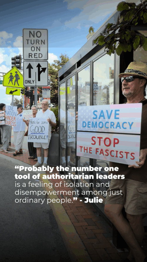 A group of people stand on a sidewalk holding protest signs, including “Save Democracy, Stop the Fascists,” “Here for some good trouble!” and “No Nation Under Duress.” One man wears sunglasses and a hat.