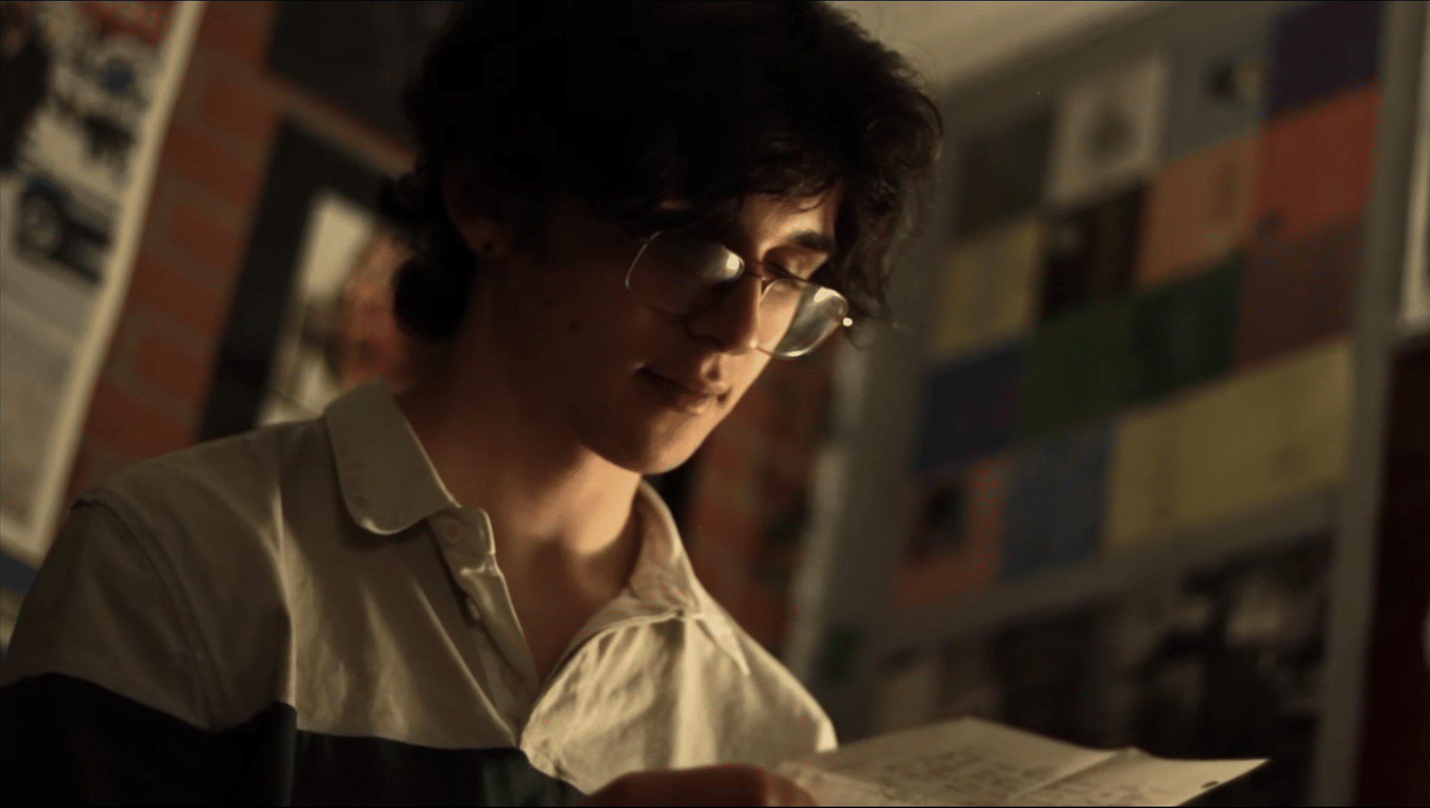 A young person with glasses and dark curly hair reads a letter in a warmly lit room, with colorful papers and posters—some from Montclair Film Festival 2025 Events—covering the wall in the background.